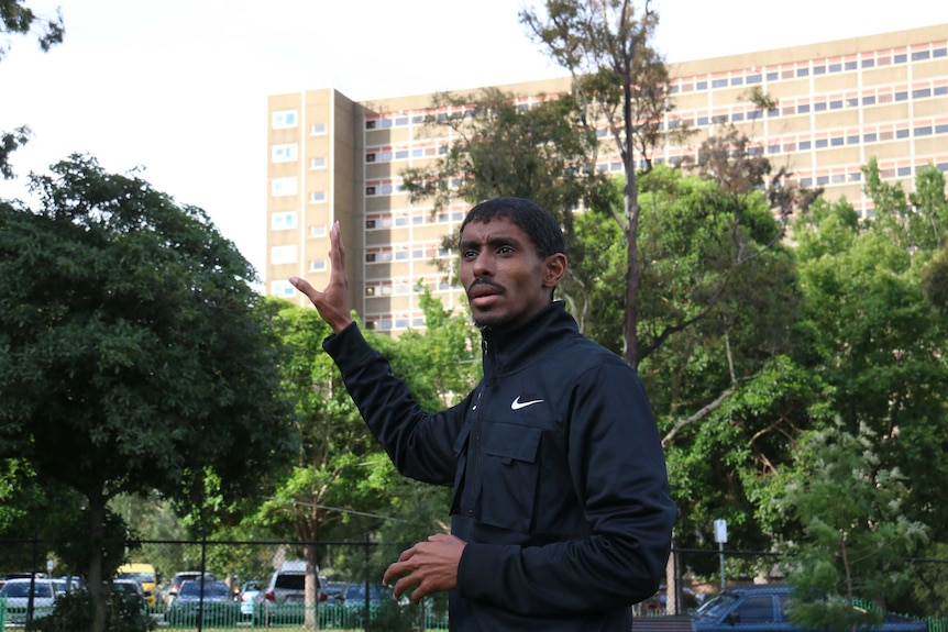 Ahmed Dini gestures with his hands on the soccer field, with the social housing towers in North Melbourne in the background.