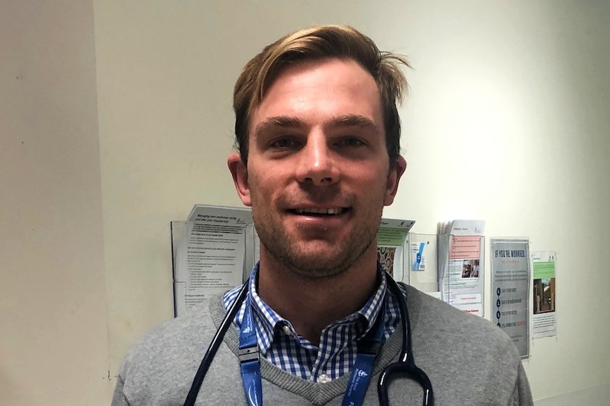 A man in his thirties smiling in a hospital corridor with a stethoscope around his neck.