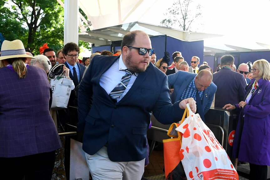 A man dressed in a suit and tie, wearing sunglasses and carrying bags, moves through the barriers at Flemington.