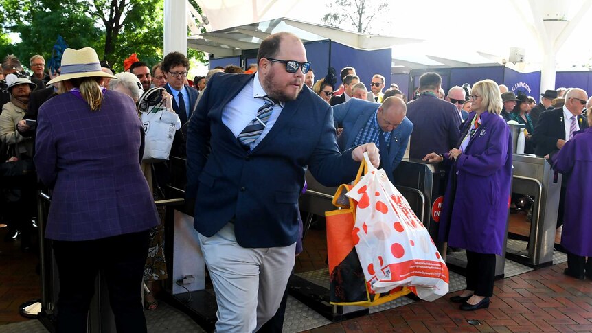 A man dressed in a suit and tie, wearing sunglasses and carrying bags, moves through the barriers at Flemington.
