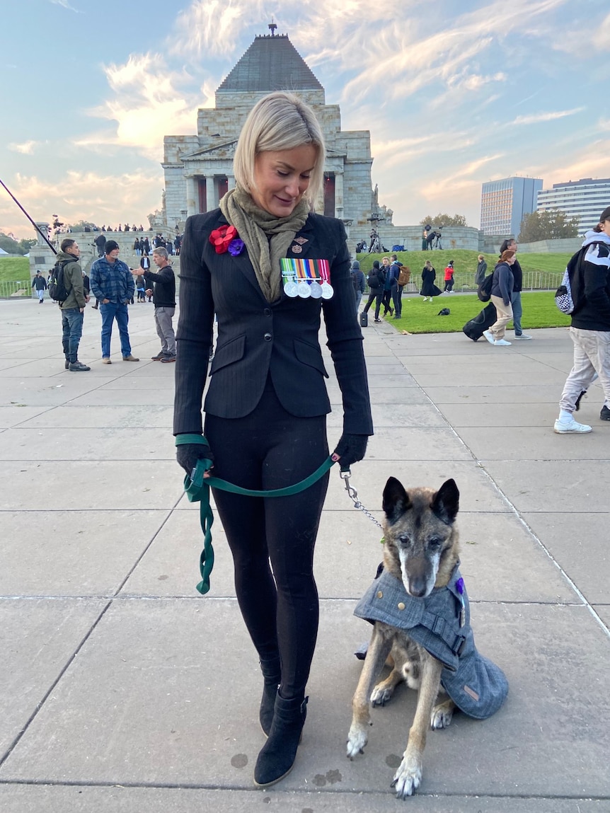 A woman with a dog wearing a coat, both with military medals and poppies, standing in front of the Shrine of Remembrance.