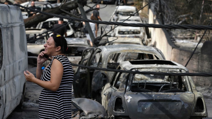 A woman speaking on a phone looks distressed with burnt-out cars in the background.