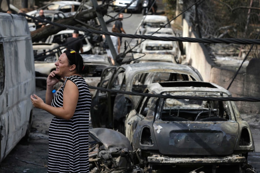 A woman speaking on a phone looks distressed with burnt-out cars in the background.