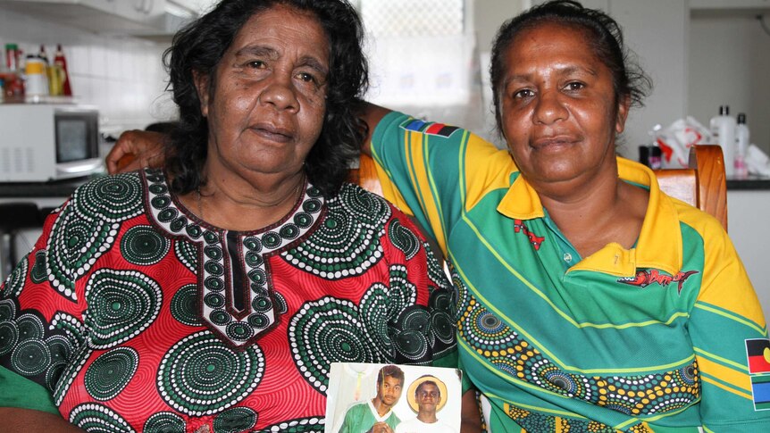 Two women sit on chairs in their home, holding a photo of two young men.