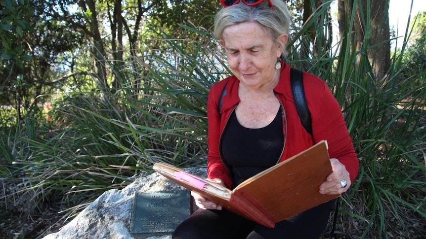 A woman in her 60s waring a red jacket sits on a rock with a plaque reading the large, old council minute book.