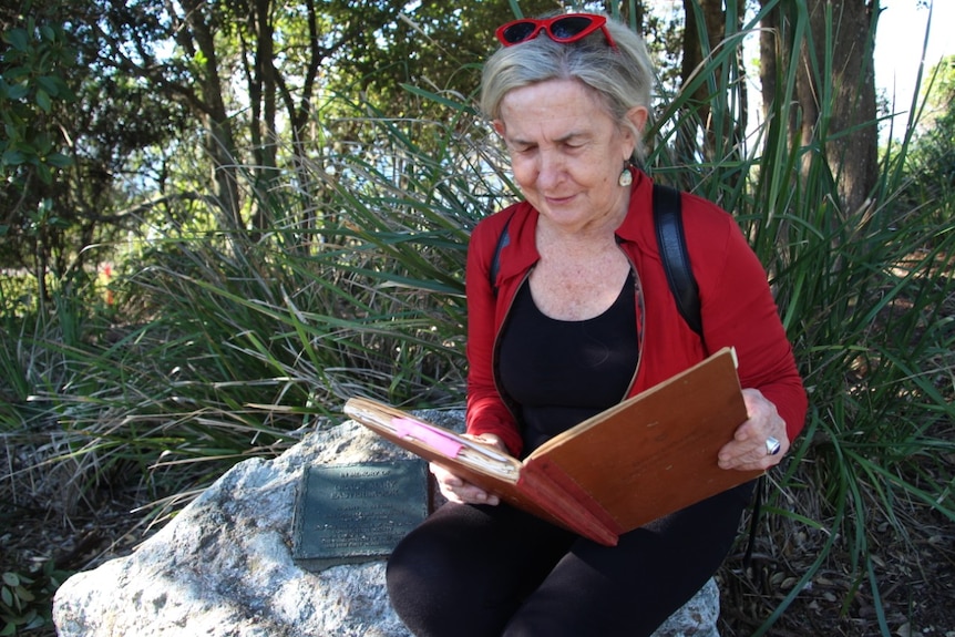 A woman in her 60s waring a red jacket sits on a rock with a plaque reading the large, old council minute book.
