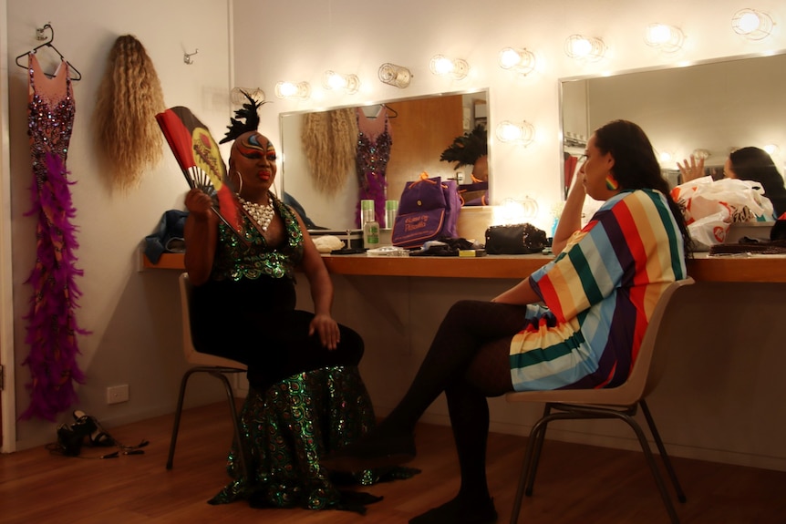 A man in drag fanning himself and a woman in a rainbow dress sit facing each other talking. 