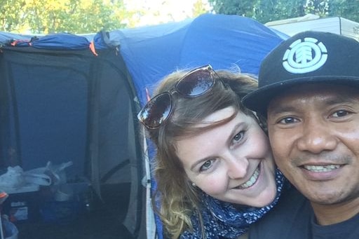 A couple smile for a selfie. There is a tent in the background.
