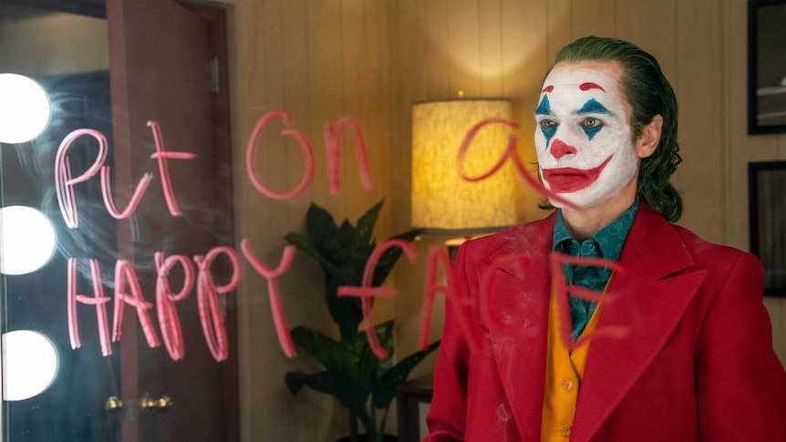 Actor Joaquin Phoenix dressed as the Joker is looking into a mirror that has the words 'put on a happy face' written on it