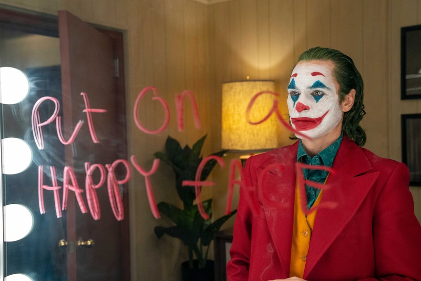 Actor Joaquin Phoenix dressed as the Joker is looking into a mirror that has the words 'put on a happy face' written on it