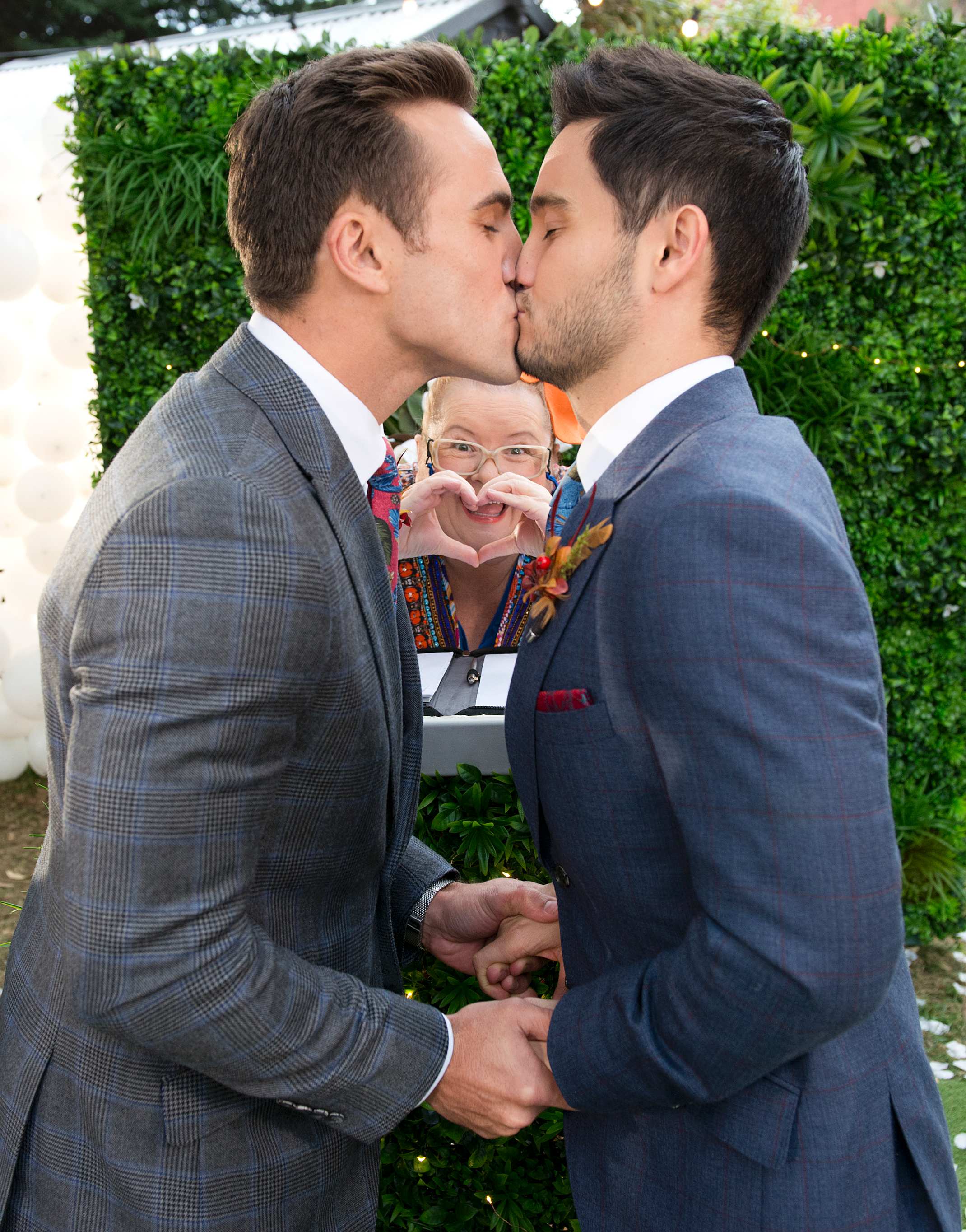 A picture of Neighbours characters Aaron and David kissing with Magda Szubanski making a heart sign.