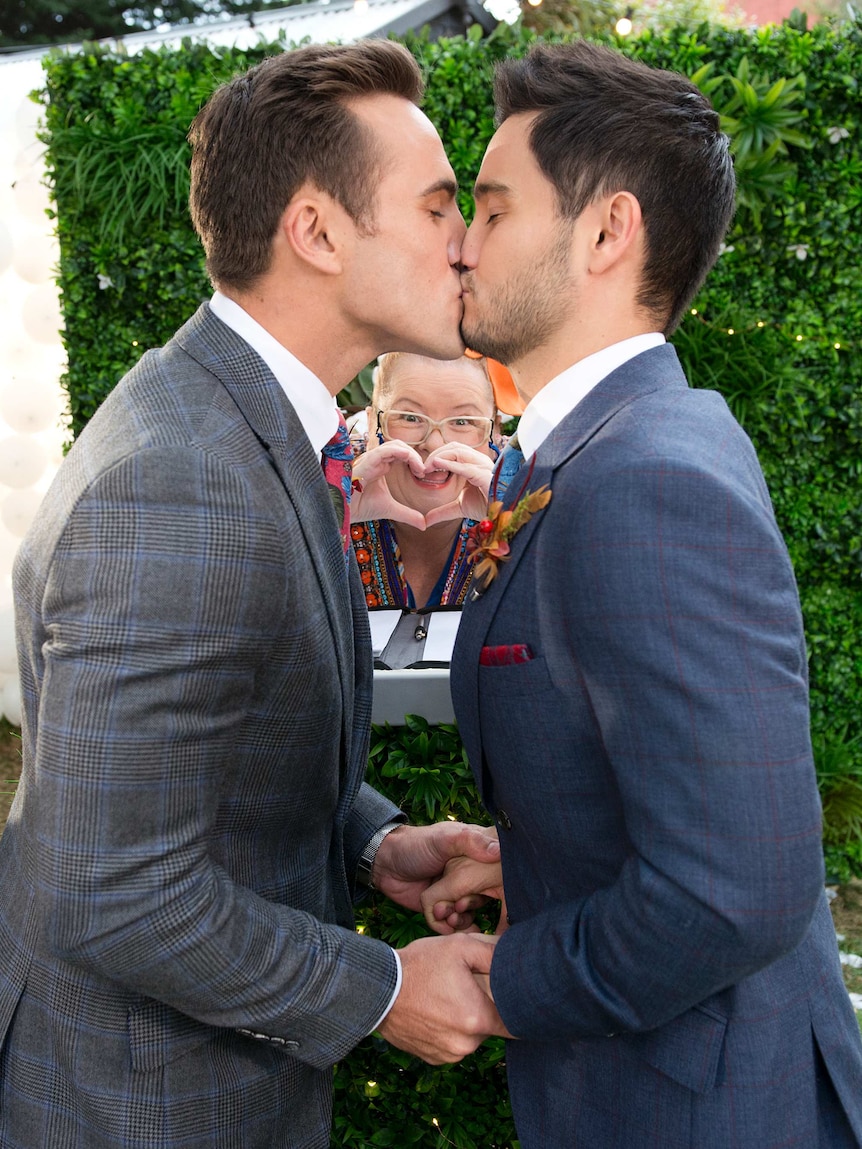 A picture of Neighbours characters Aaron and David kissing with Magda Szubanski making a heart sign.