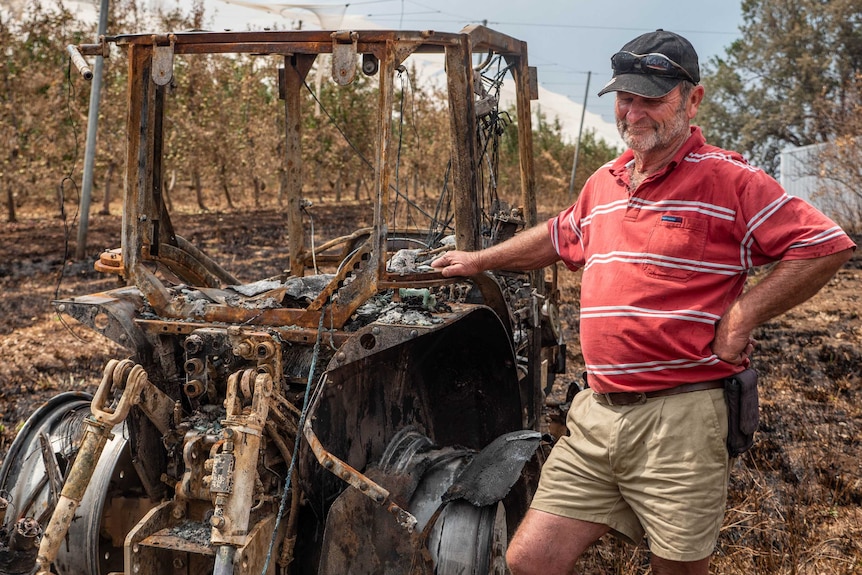 Greg Mouat wears a red t-shirt, shorts and cap and looks despondently at a burnt out piece of machinery.