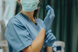A picture of a woman in scrubs, cant see her face except that she's wearing a face mask and putting gloves on hands