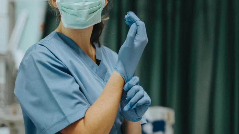 A picture of a woman in scrubs, cant see her face except that she's wearing a face mask and putting gloves on hands