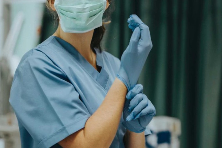 A picture of a woman in scrubs, cant see her face except that she's wearing a face mask and putting gloves on hands