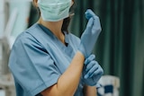 A picture of a woman in scrubs, cant see her face except that she's wearing a face mask and putting gloves on hands