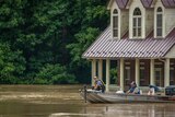 Two mean in a tin boat float past a brick colonial home partially submerged by brown floodwaters.