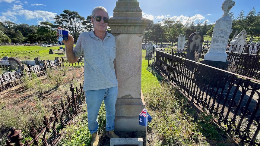 David Potts stands next to memorial statue wearing grey shirt and sunglasses, holding war medals. 