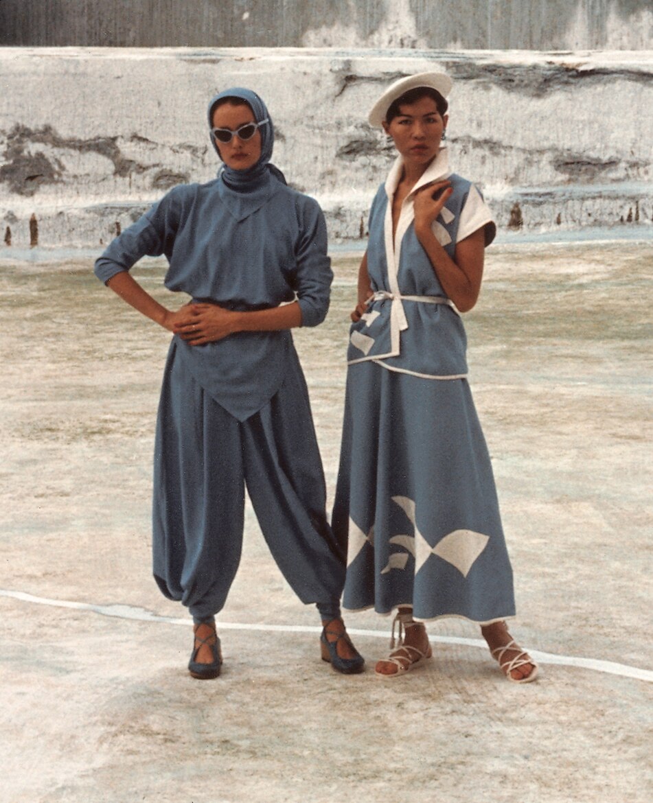 One woman wearing blue top and pants and second woman wearing dress with white shapes like the Opera House.