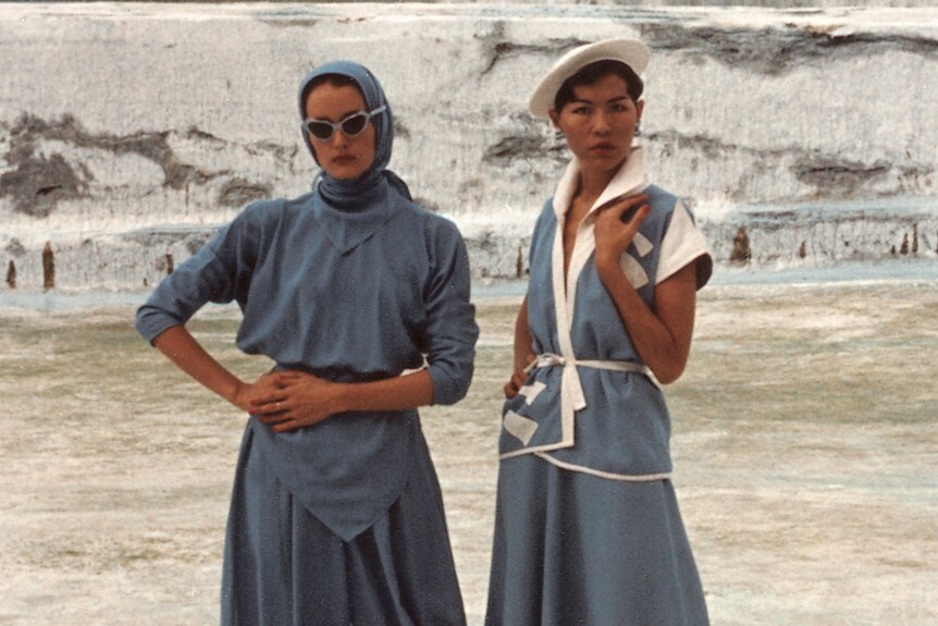 One woman wearing blue top and pants and second woman wearing dress with white shapes like the Opera House.