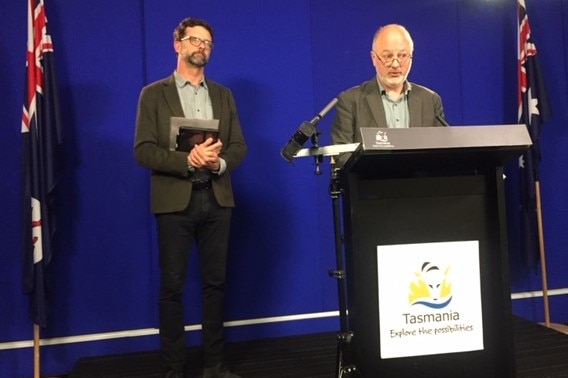 A bald man with a beard and glasses stands at a podium, another taller, bearded man stands behind him holding paperwork.