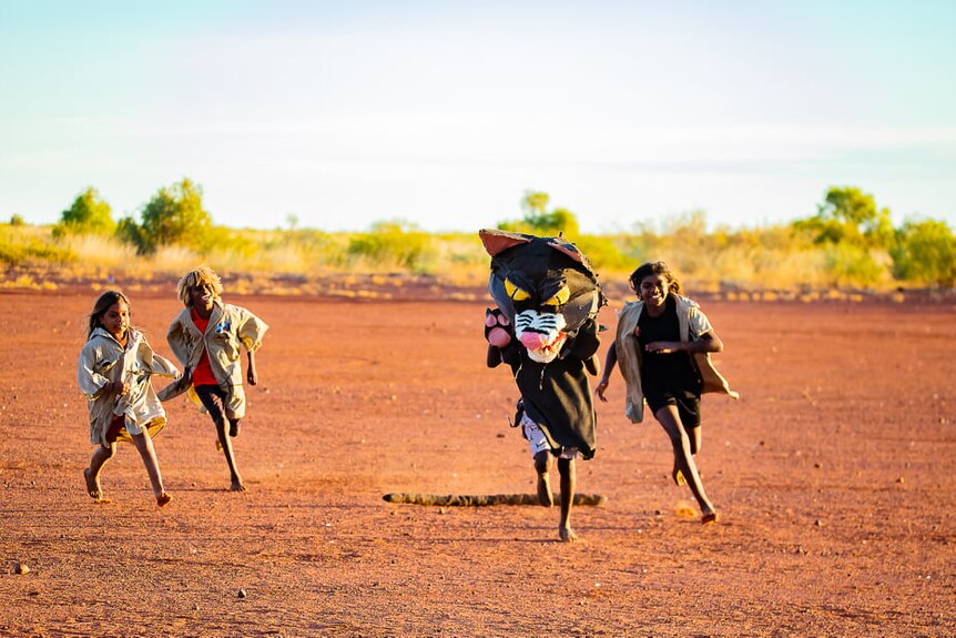a child wearing a cat head is chased across the red dirt by three children