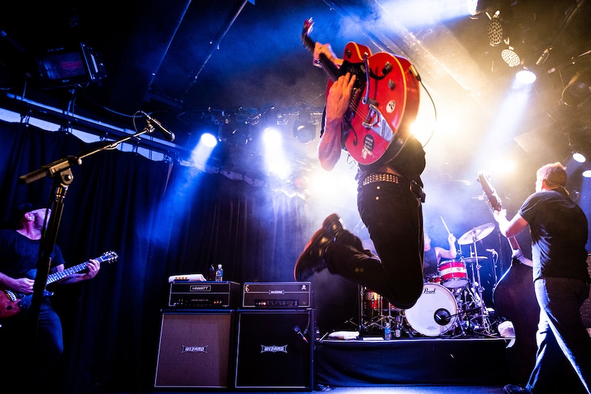 A guitar player on a stage jumps in the air with bandmates nearby