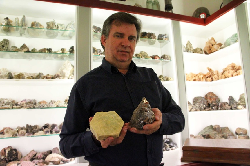 A man holds two large mineral specimens, one yellow with a smooth surface, the other shiny, silver, made of many smaller prisms.