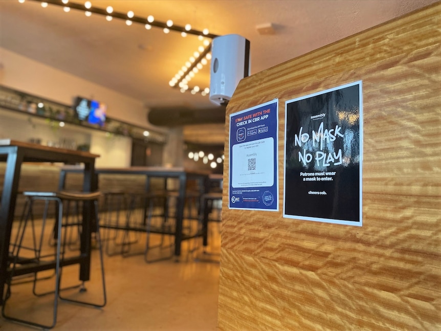 The front desk of a bar, with a sanitiser dispenser, a QR code for the Check In CBR app and a sign about wearing masks.