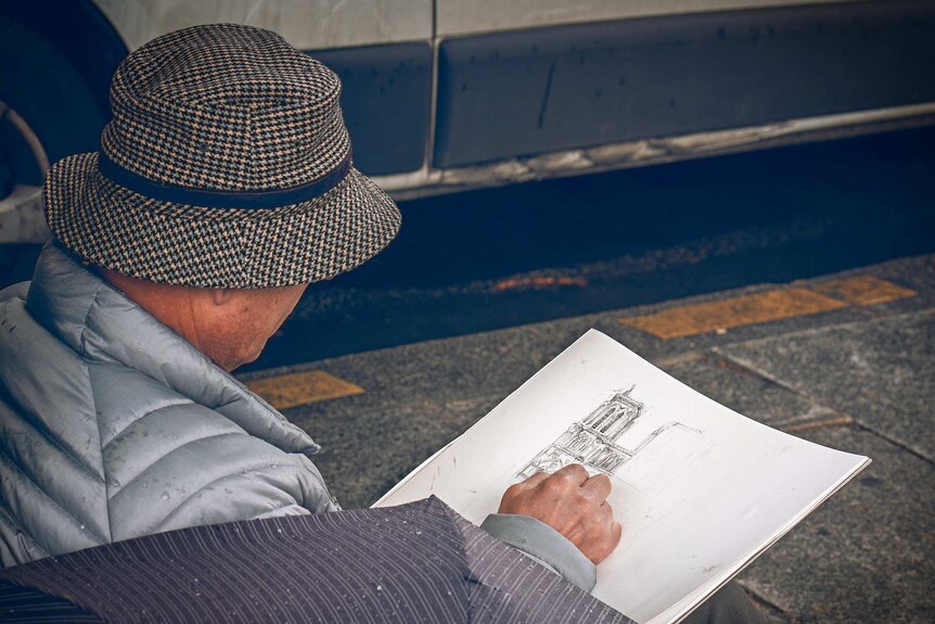 A man wearing a checked hat and puffy jacket sketches half of Notre Dame cathedral in pencil.