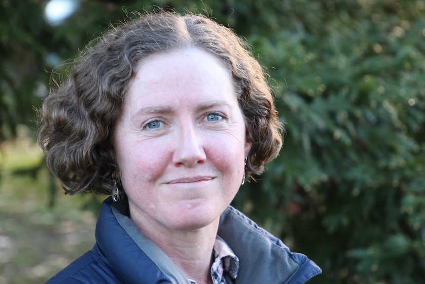 Head and shoulders portrait of a woman smiling, standing in front of greenery in the outdoors.