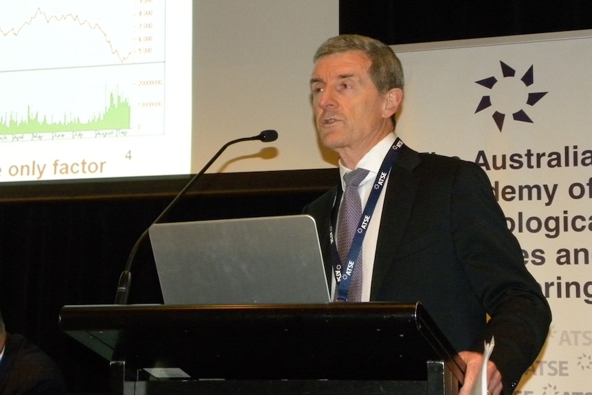 A grey-haired man on the podium addressing a conference. He is wearing a dark suit, grey tie and white shirt