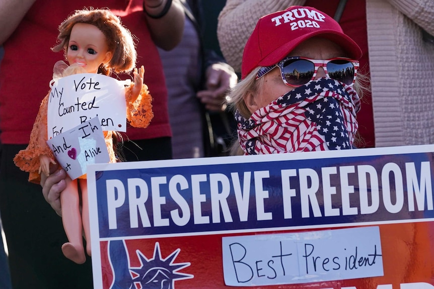 A woman wearing a Trump 2020 red cap and American flag glasses and mask holds a doll saying My vote counted for Biden.