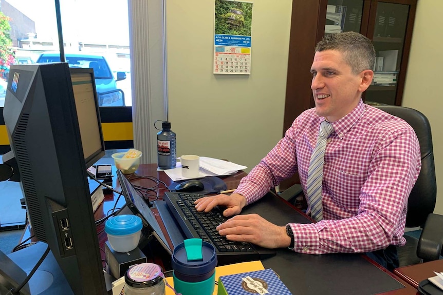 A man in a checked shirt and tie sitting at his desk typing on a desktop computer.