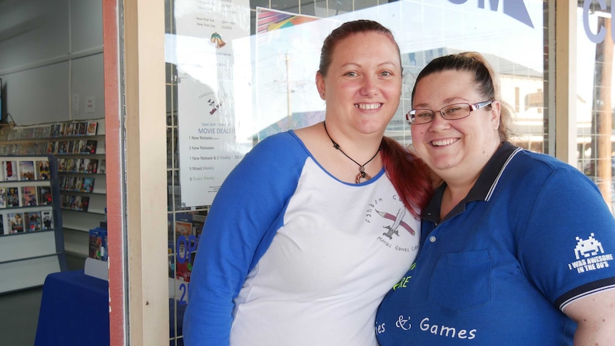 Two women face the camera and smile outside a video rental shop, with shelves of movies visible through the front door.