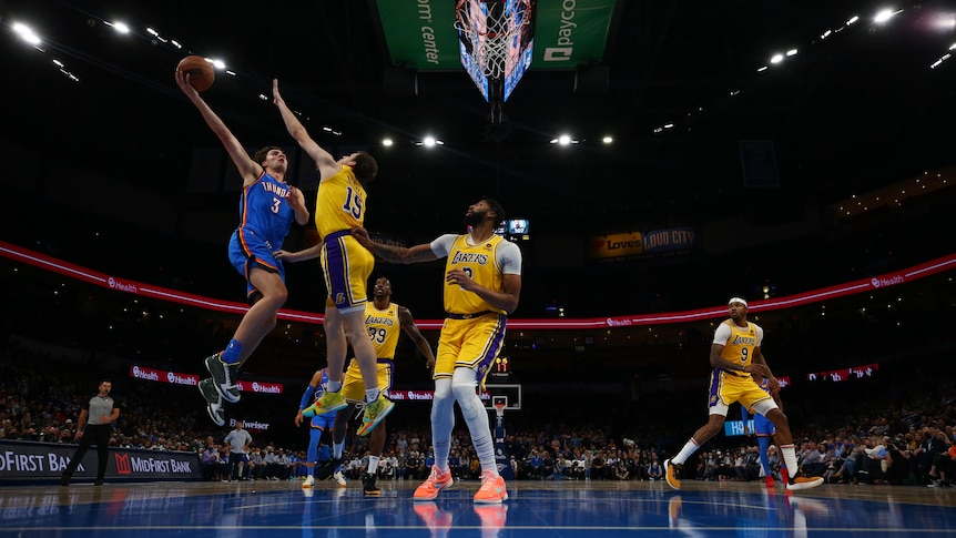 An Australian NBA player jumps in the air and tries to release a hook shot as a defender tries to block him.