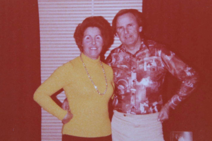 Two people stand in front of venetian blinds in 1980s clothing