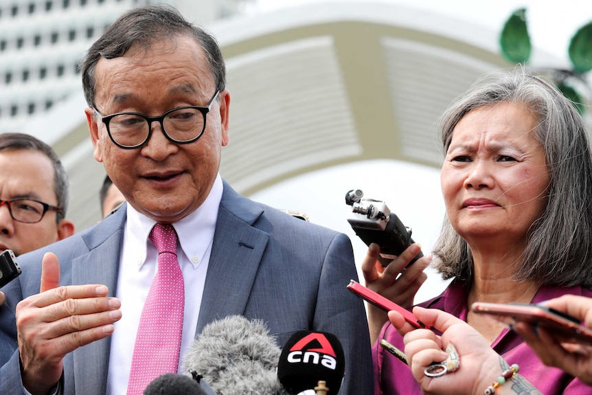 Sam Rainsy gestures as he speaks in front of press microphones and Mu Sochua stands stony-faced beside him.