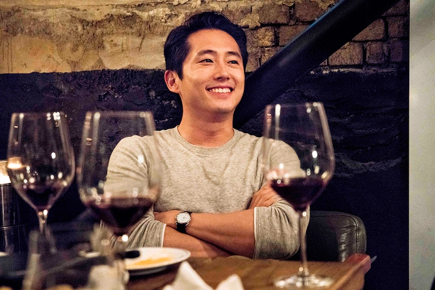 Colour still of Steven Yeun smiling in front of table of wine glasses 2018 film Burning.