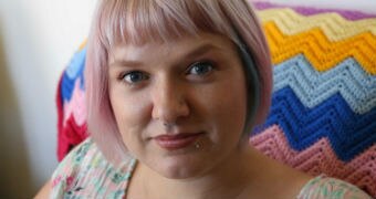 A young woman with pink-blonde hair and blue eyes look sits on a couch in her home.