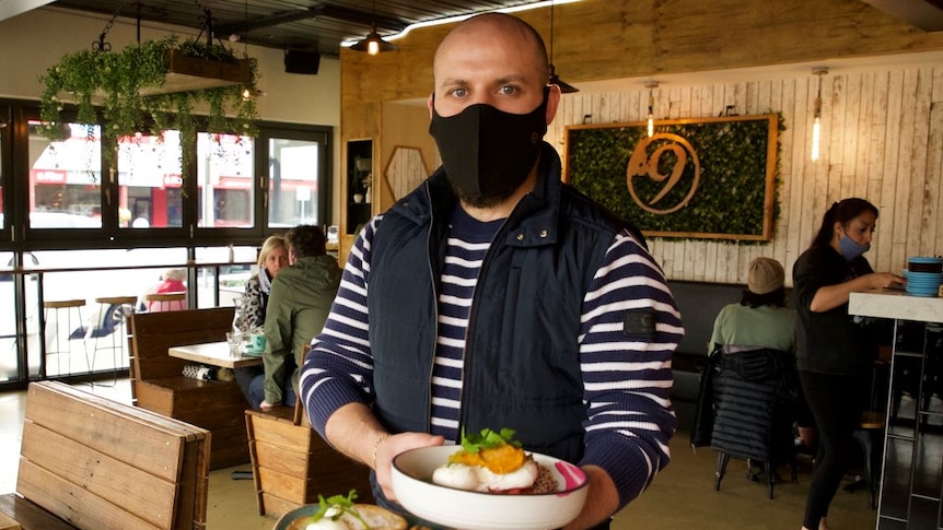 A man stands in a cafe wearing a facemask, blue and white striped jumper, holding two plates filled with bacon, eggs and toast.