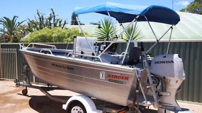 A Stacer boat with a Honda Motor, and a blue canopy, sitting on a boat trailer in a carpark on a sunny day.