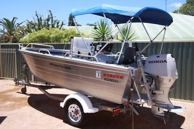 A Stacer boat with a Honda Motor, and a blue canopy, sitting on a boat trailer in a carpark on a sunny day.