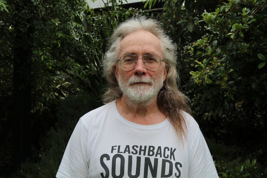 An older man with a silver beard and long grey hair sits in a garden wearing a shirt that says flashback sounds.