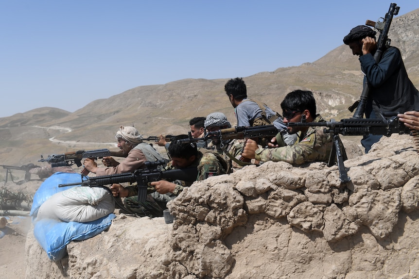 A group of men fire weapons from behind a sandbag wall