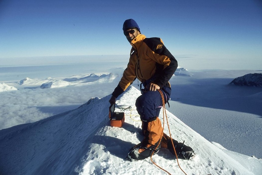 At the top of one of a series of huge, snow-capped mountains, Damien Gildea, in snow clothes, beanie and sunglasses, smiles.