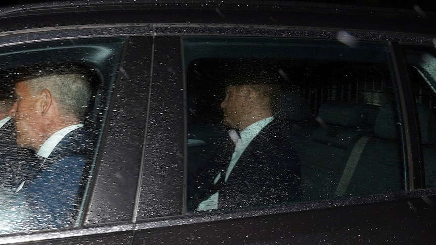 Prince Harry is pictured in the back of a black car. Rain droplets cover the window.