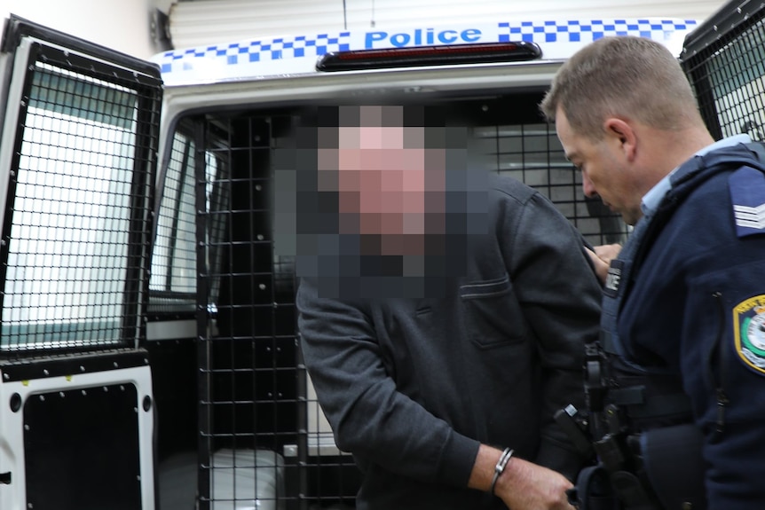 A man whose face  is heavily blurred is led out of a paddy wagon in handcuffs by a police officer.