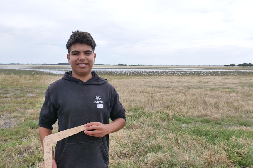 A young Aboriginal man holding a boomerang standing in front of a wetland. 